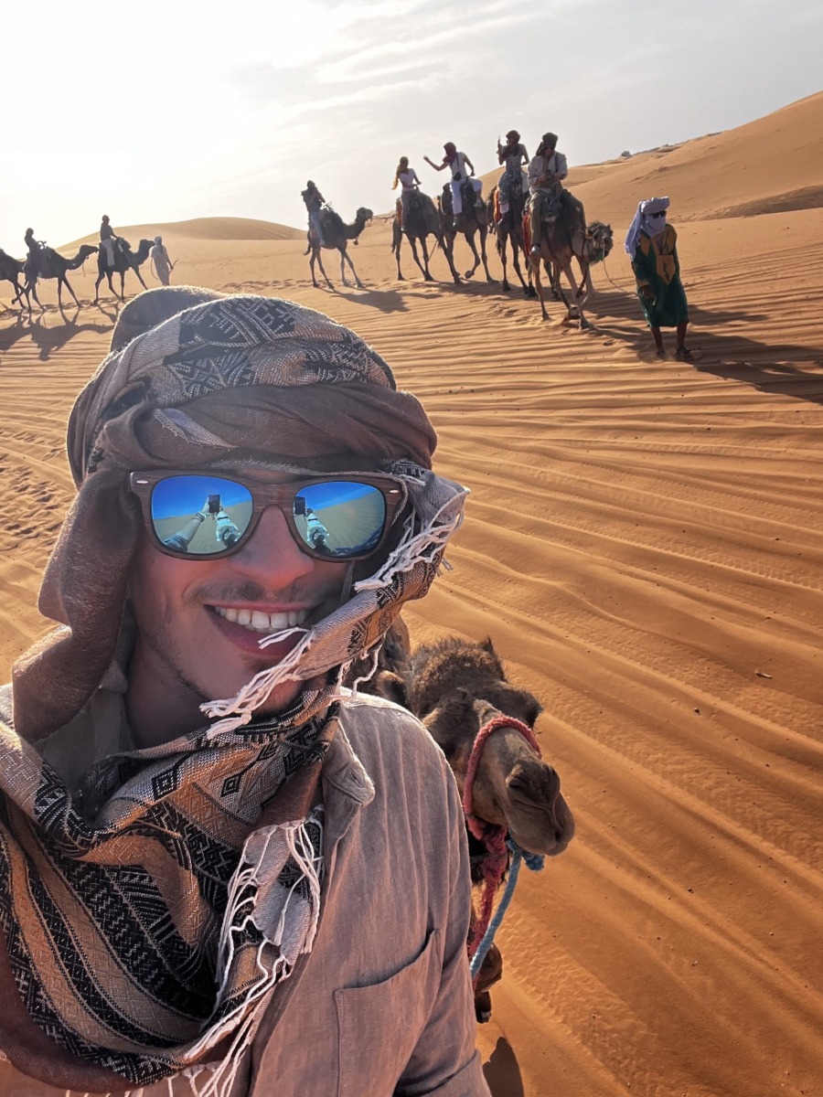 A selfie of Alex standing among the sand dunes in the Merzouga Desert in Morocco with a line of camel riders in the background.